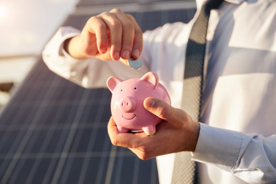 Crop unrecognizable businessman inserting coin into piggy bank while standing against blurred photovoltaic solar panel as concept of cheap alternative energy